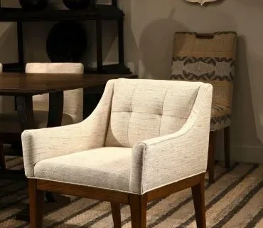 A light-colored, tufted armchair is shown in a room with a wooden table and another chair in the background.